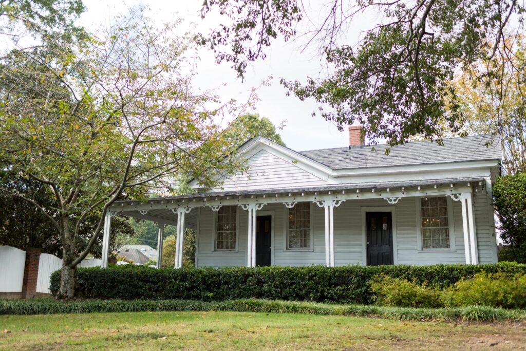 A small, light gray house with a covered front porch, white trim, and two black front doors stands behind a row of bushes and a large tree—an inviting spot often chosen for intimate event venues—with green grass and a cloudy sky in the background.