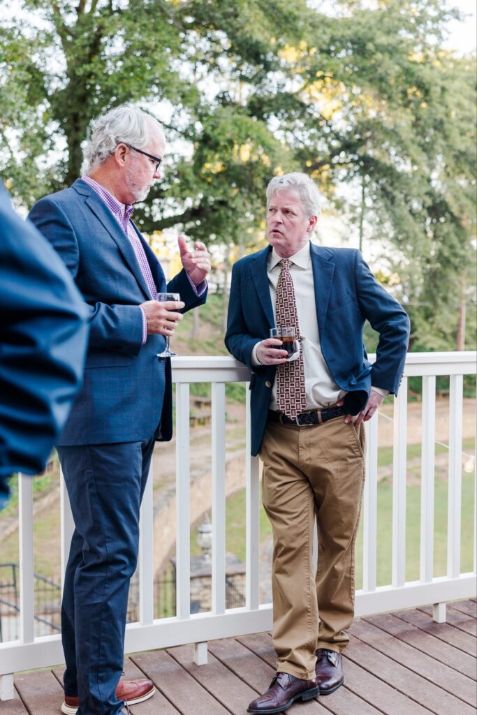 Two men in suits stand on a wooden deck at one of the event venues near Decatur, holding drinks and having a conversation. Trees and a grassy area are visible in the background as one man gestures while speaking.