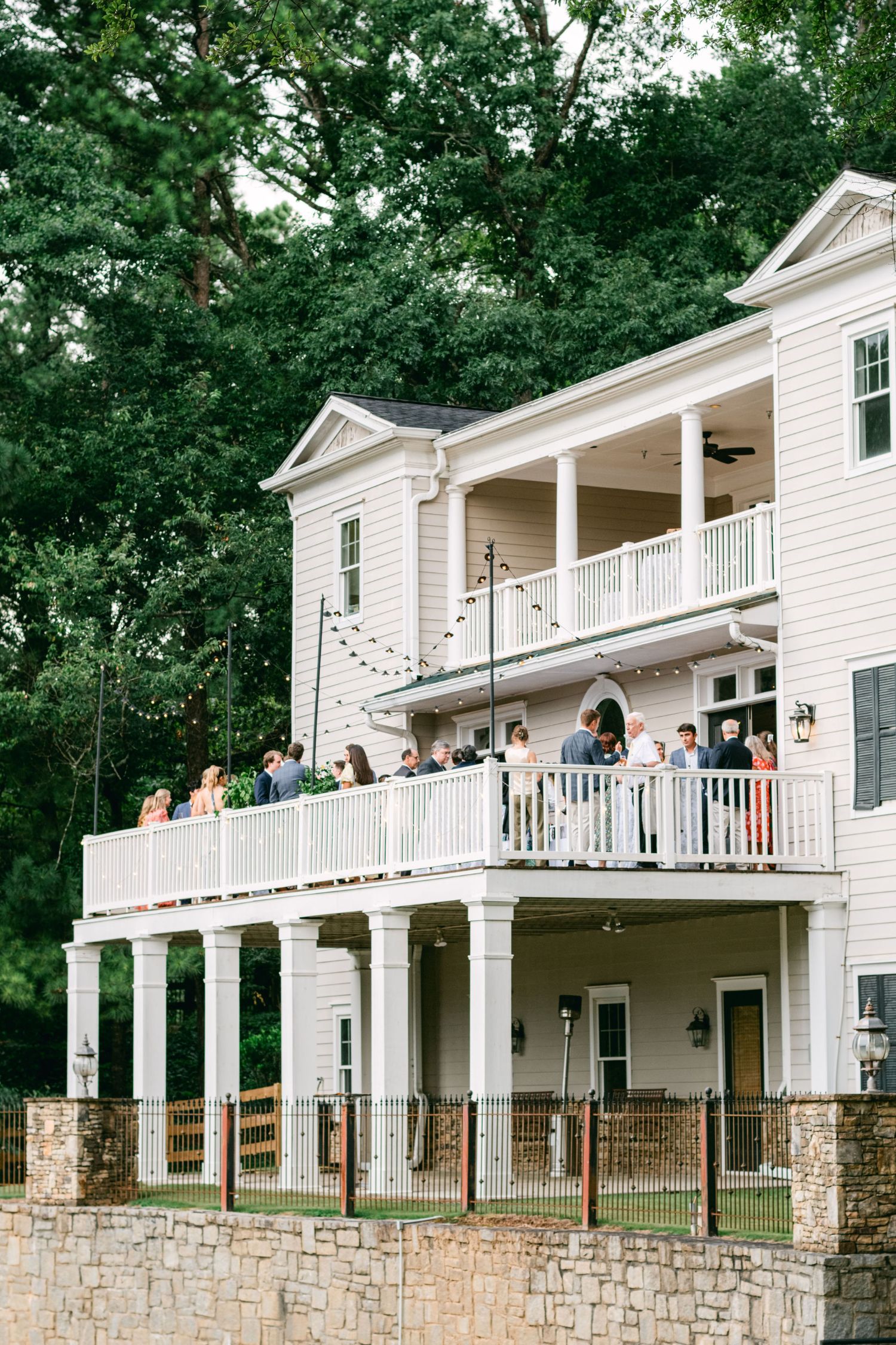 A group gathers on the upper balcony of a grand white house in Atlanta for a rehearsal dinner, surrounded by trees and a stone wall. String lights twinkle overhead, setting the scene for an unforgettable southern rehearsal dinner menu.