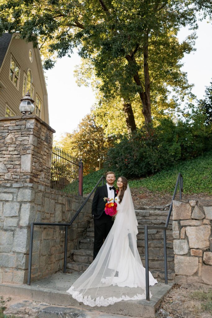 A bride and groom stand together on stone steps outdoors at their Chastain Horse Park wedding reception, surrounded by greenery. The bride wears a long white gown and holds a bright bouquet as sunlight filters through the trees above.
