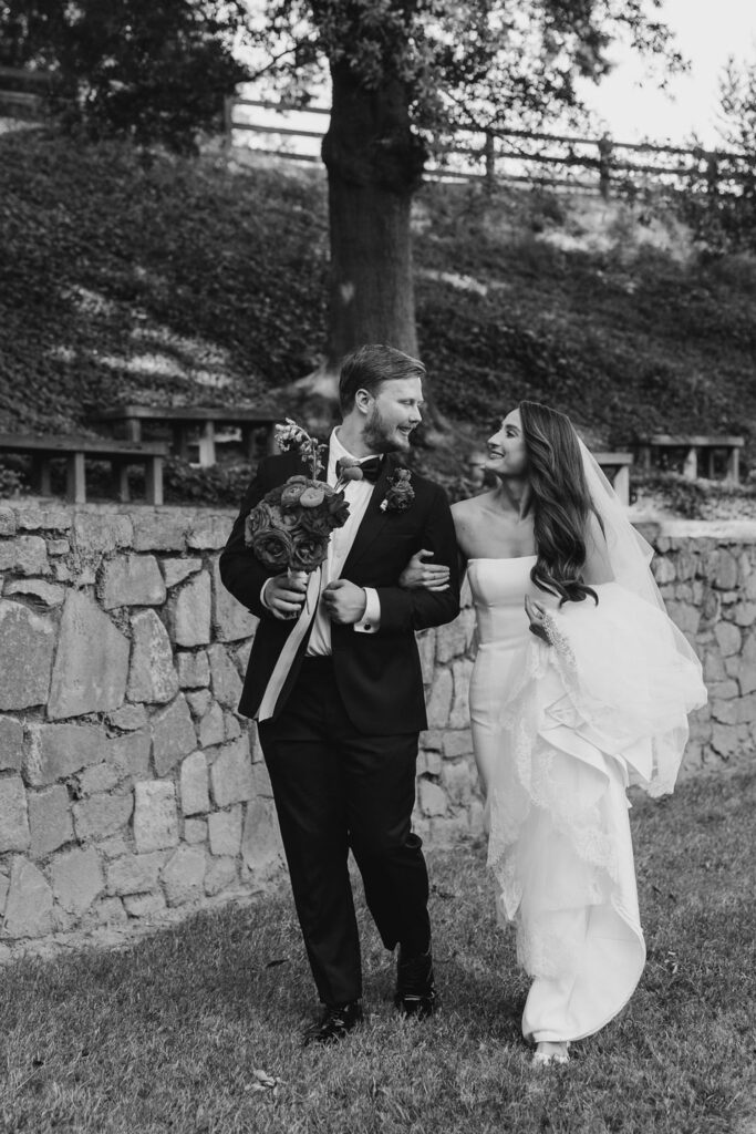 A bride and groom walk arm in arm outdoors, smiling at each other in front of a stone wall with trees beyond—captured just before their Chastain Horse Park wedding reception. The groom holds a bouquet; the bride wears a strapless gown and veil.