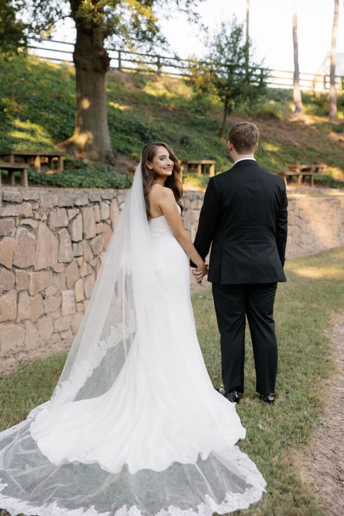 A bride in a white gown and long veil holds hands with a groom in a black suit as they stand outdoors on grass near a stone wall during their Chastain Horse Park wedding reception. The bride looks back and smiles while the groom faces away.