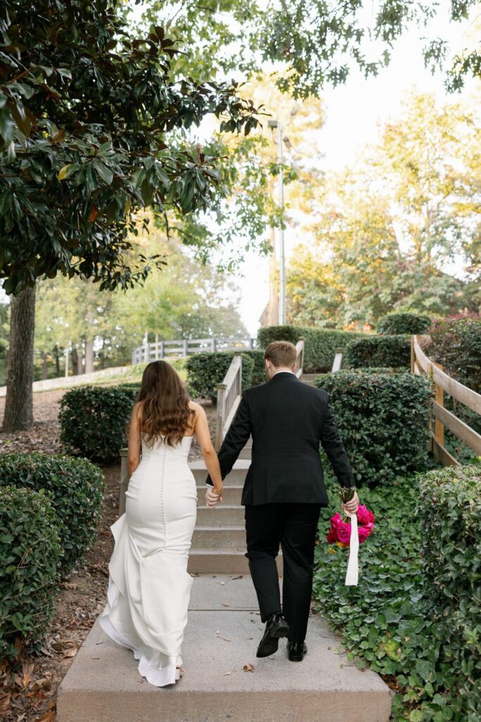 A bride in a white dress and a groom in a black suit walk up outdoor steps at their Chastain Horse Park wedding reception, holding hands. The groom carries bright pink flowers as green bushes and trees line the walkway under a sunny sky.