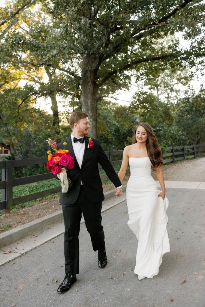 A bride in a white dress and a groom in a black tuxedo holding a colorful bouquet walk hand in hand on a paved path at their Chastain Horse Park wedding reception, smiling with trees and a black fence in the background.