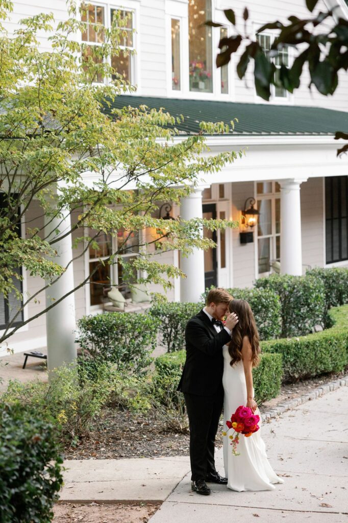 A groom in a black suit gently kisses the forehead of his bride, who holds a bright pink bouquet. They stand close together on a sidewalk in front of a white building with greenery after their Chastain Horse Park wedding reception.