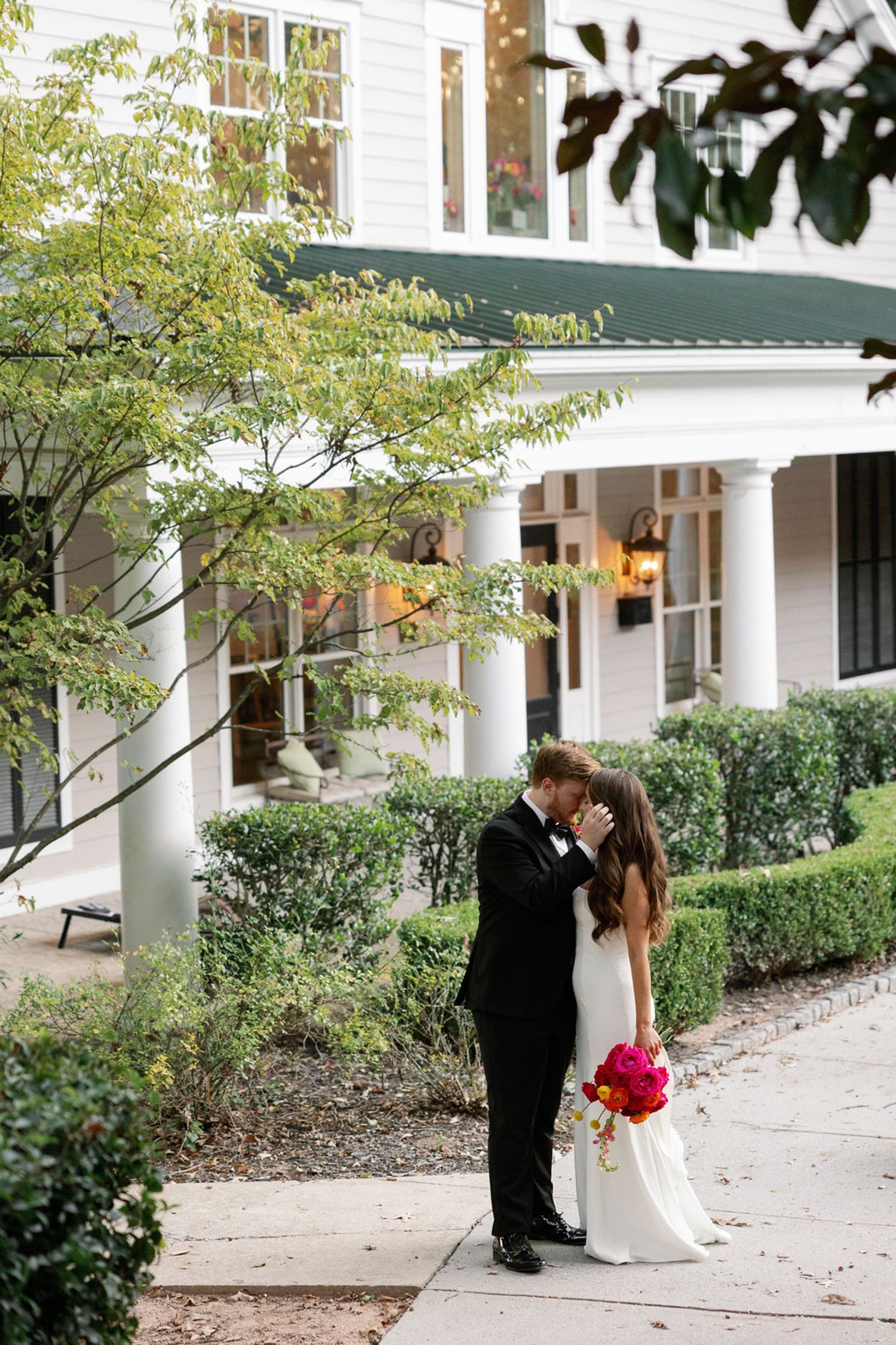 A groom in a black suit gently kisses the forehead of his bride, who holds a bright pink bouquet. They stand close together on a sidewalk in front of a white building with greenery after their Chastain Horse Park wedding reception.