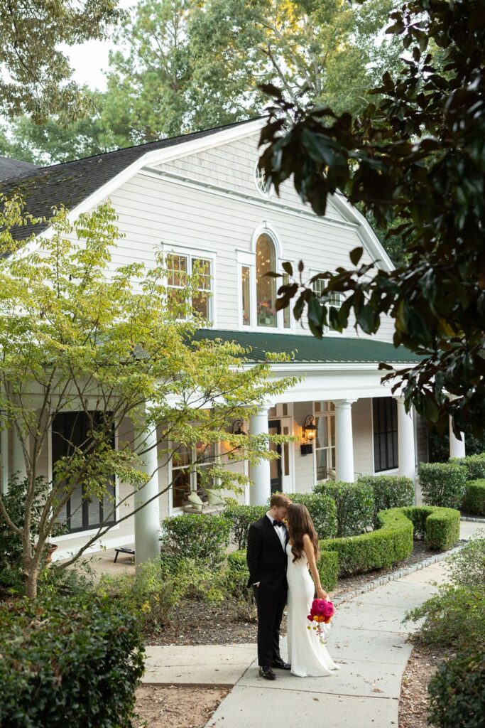 A bride and groom stand closely together on a curved sidewalk, surrounded by greenery, in front of a large, light-colored house with columns and tall windows—captured just before their Chastain Horse Park wedding reception. The bride holds a bright bouquet.