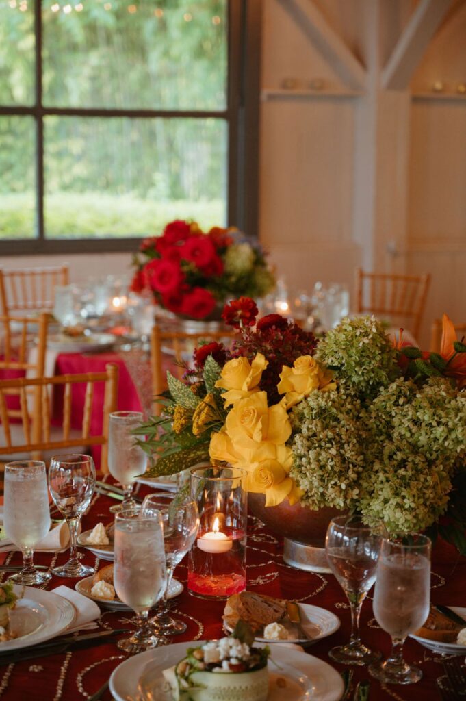 A beautifully set banquet table for a Pace House wedding reception features elegant glassware, plates, water glasses, and lit candles. Large floral centerpieces with yellow, red, and green blooms complement the wooden chairs and window view.