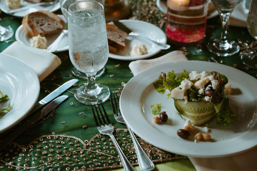 A close-up of a Pace House wedding reception dinner table set with a glass of water, plates of bread with butter, and a salad with olives, cheese, lettuce, and cucumbers on a white plate atop a green, decorated tablecloth.