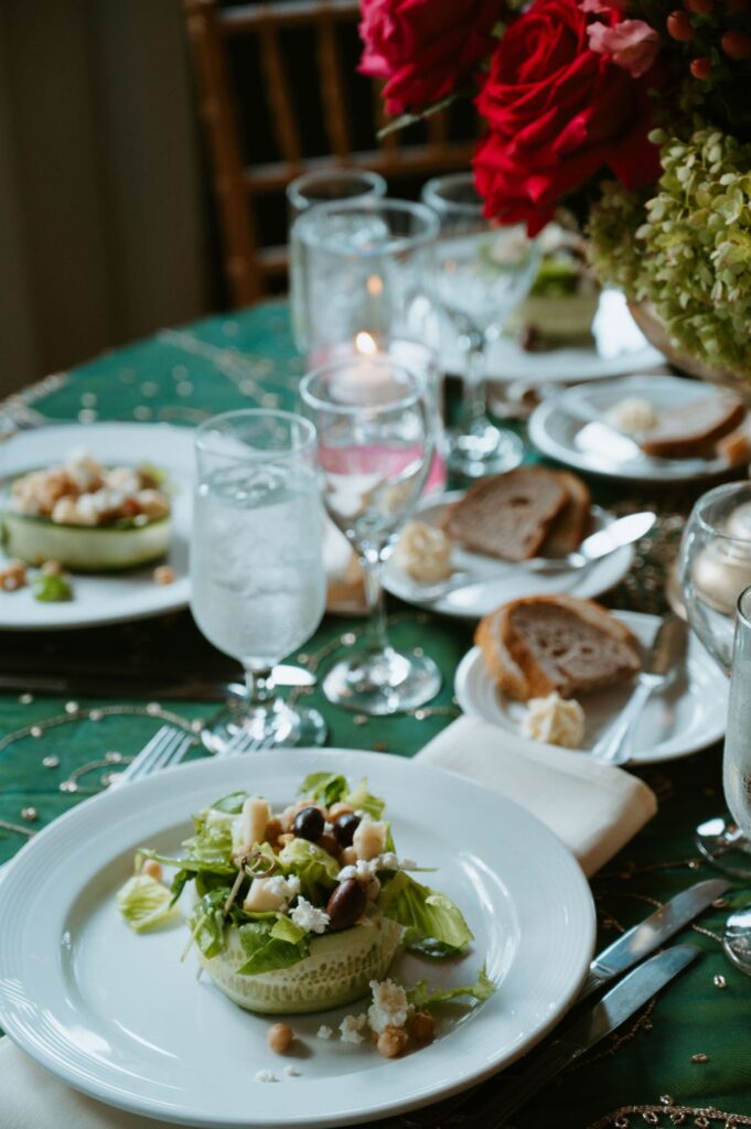A round salad topped with lettuce and chickpeas sits on a white plate at a Pace House wedding reception, set on a green tablecloth. Slices of bread, water glasses, and a flower arrangement complete the elegant table setting.