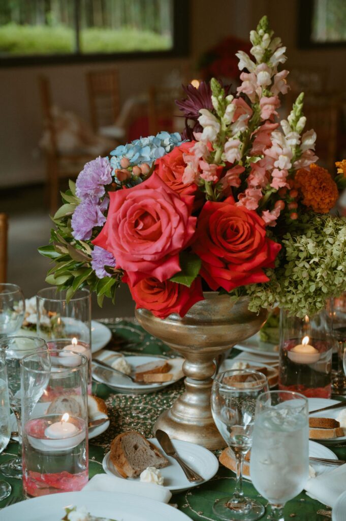 A gold vase filled with vibrant flowers, including red roses, pink snapdragons, and purple blooms, sits on a table set for a Pace House wedding reception with glassware, plates, bread, and floating candles, creating an elegant dining scene.