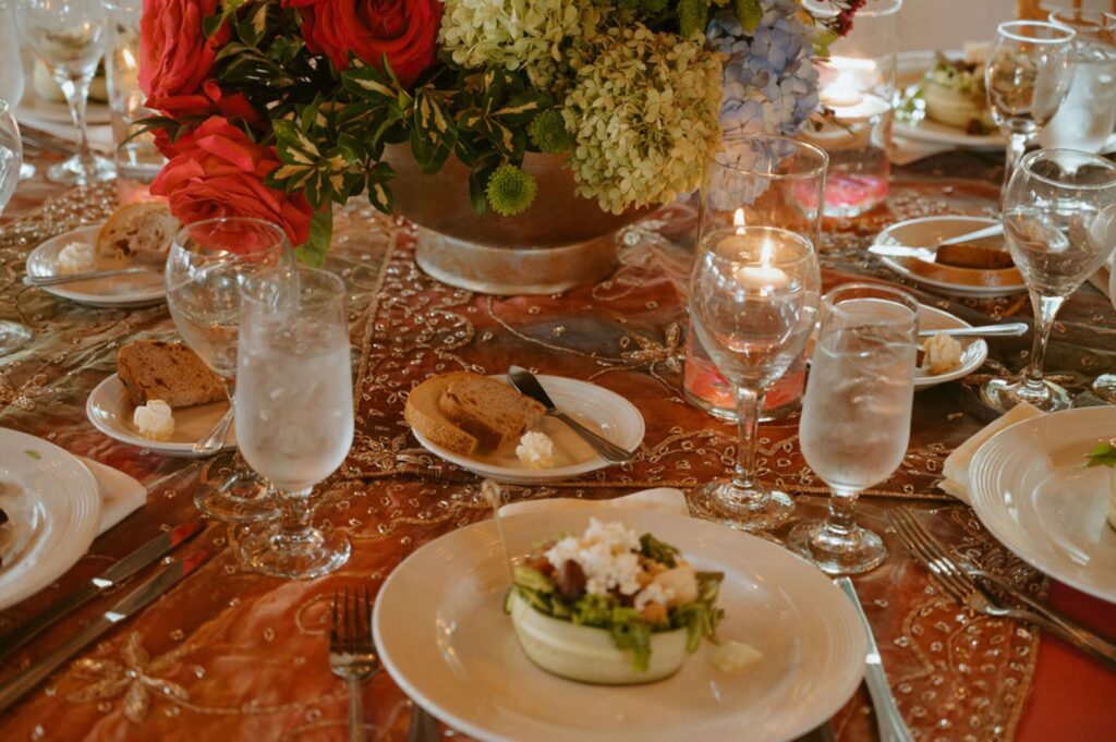An elegant Pace House wedding reception table setting features plates of salad, bread, water and wine glasses, lit candles, and a large floral centerpiece with red, white, and green flowers on a decorated tablecloth.