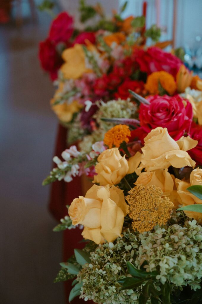 A vibrant floral arrangement featuring yellow roses, orange and red flowers, and green foliage graces the table at a Pace House wedding reception, set against a softly blurred background.