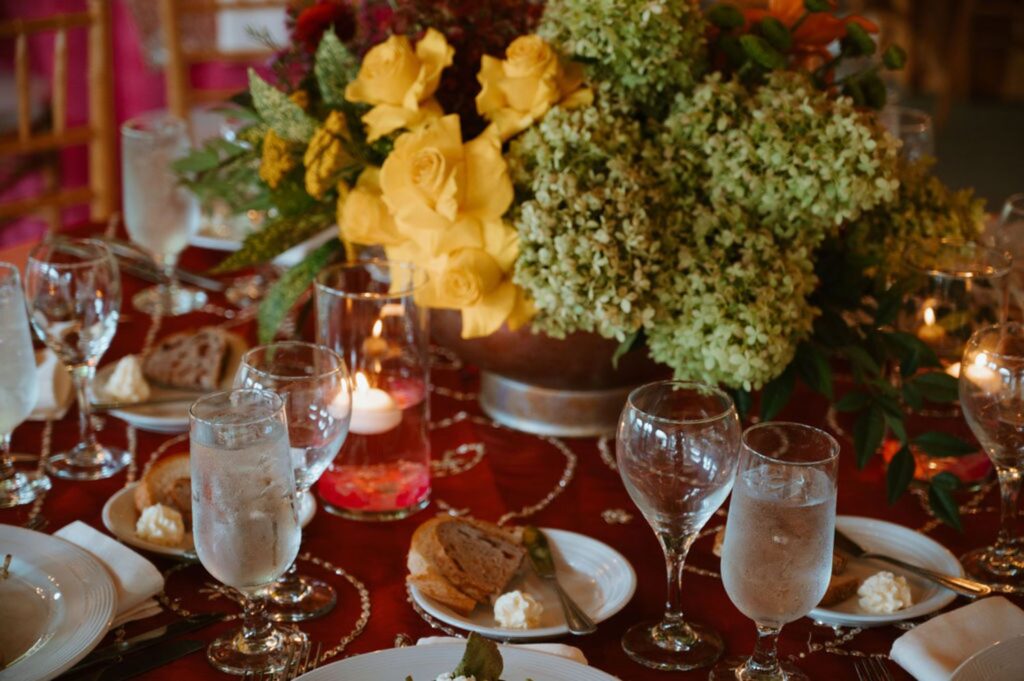 A formal dining table at a Pace House wedding reception is set with glasses of water, plates with bread and butter, silverware, and a centerpiece of yellow roses and green flowers. Lit candles in glass holders add a warm ambiance.