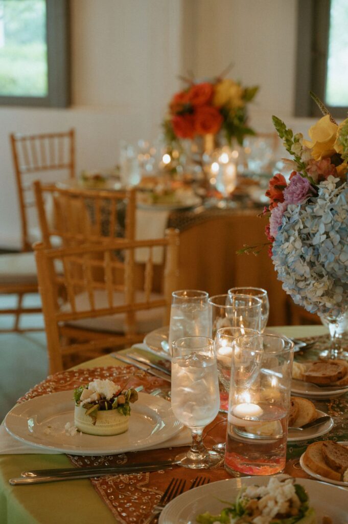 A beautifully set dining table with elegant glassware, plates of salad, lit candles, and vibrant flowers—perfect for a Pace House wedding reception. Wooden chairs surround the scene in a softly lit room with blue hydrangeas and mixed blooms.