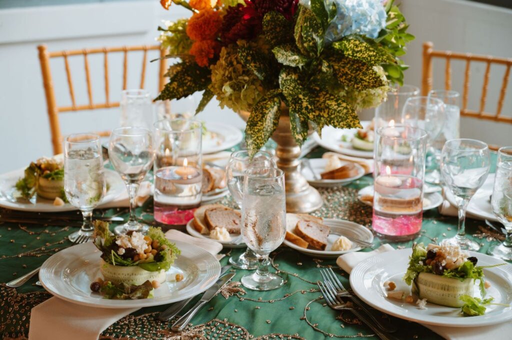A Pace House wedding reception features a formal dining table set with plates of salad, glasses of water and wine, bread slices, candles, and a large floral centerpiece on a green tablecloth with gold accents.