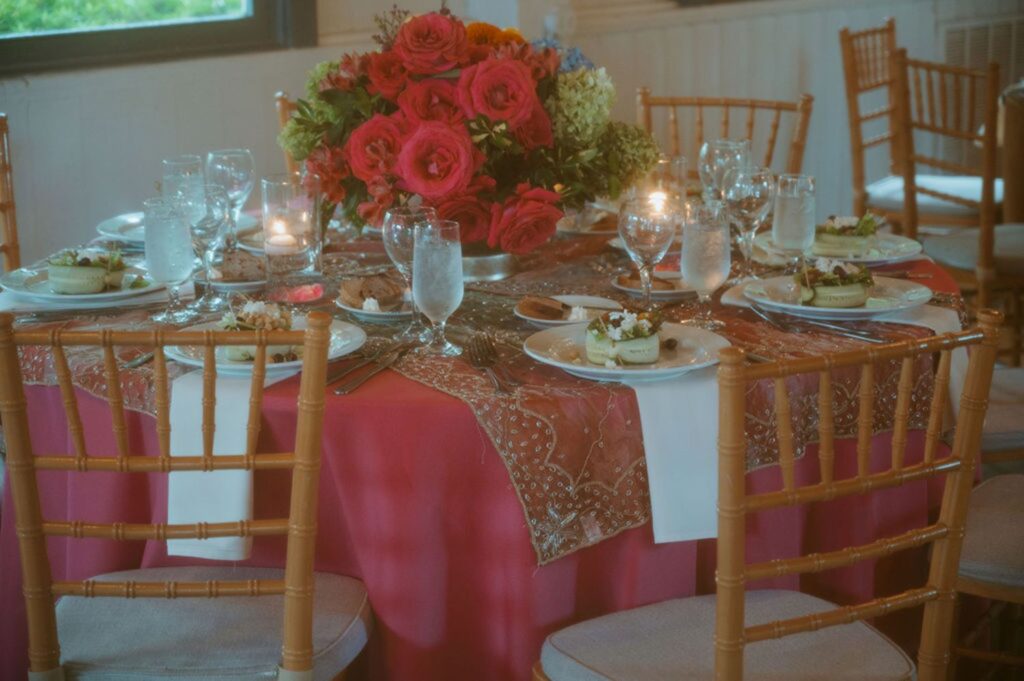 A round table set for four with wooden chairs, decorated with a bright red tablecloth and a floral centerpiece—perfect for a Pace House wedding reception—with lit candles, glasses of water, and plates of food in an elegant indoor setting.