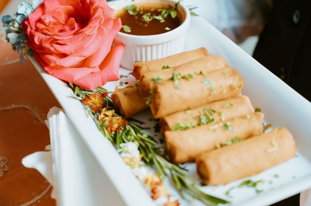 A rectangular white plate with six golden-brown spring rolls, a small cup of dipping sauce, garnished with chopped herbs, a pink rose, and assorted greens—perfect for your Pace House wedding reception.