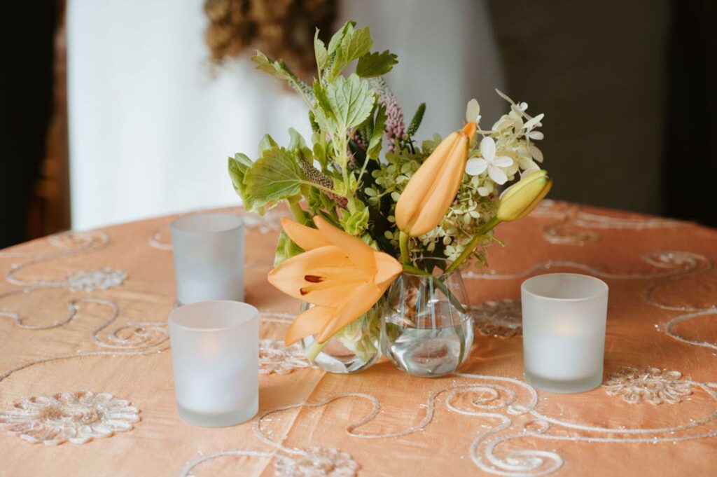 A small glass vase with orange lilies and greenery sits on a peach-colored embroidered tablecloth, surrounded by four frosted glass votive candle holders—perfect decor for an elegant Pace House wedding reception.