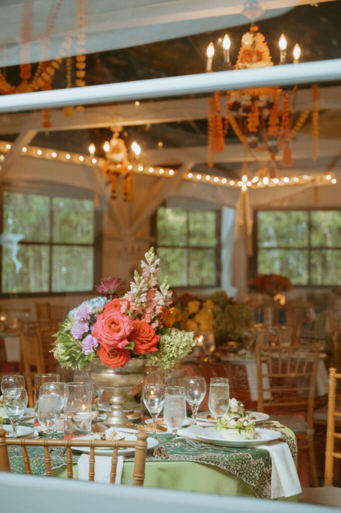 A decorated banquet hall seen through a window, set for a Pace House wedding reception with plates, glassware, and a centerpiece of pink, red, and purple flowers beneath chandeliers and string lights overhead.