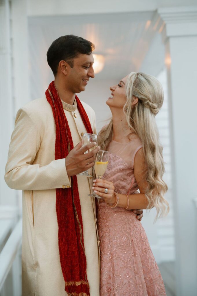 A man in a cream sherwani with a red scarf and a woman in a pink dress smile at each other, holding glasses of champagne, standing closely together at their elegant Pace House wedding reception.