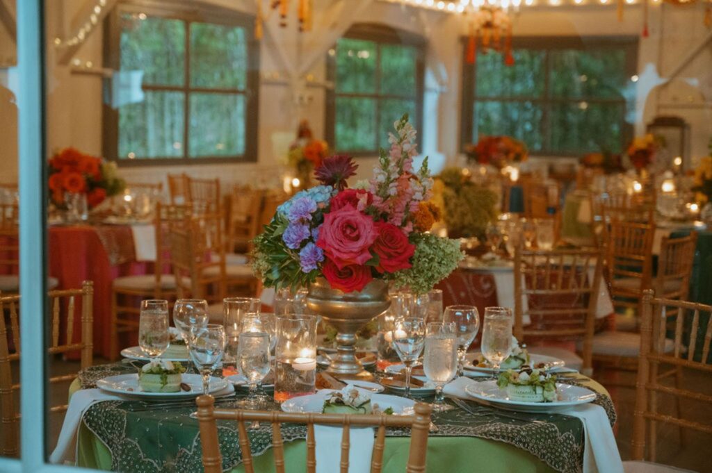 A decorated banquet table with a vibrant floral centerpiece, surrounded by neatly arranged plates, glasses, and candles sets the scene for a Pace House wedding reception in an elegant, warmly lit room with large windows and wooden chairs.