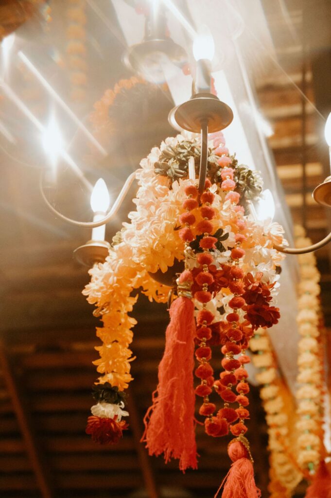 A chandelier decorated with orange, yellow, and red marigold garlands, flowers, and tassels glows warmly at a Pace House wedding reception in a festive indoor setting with strings of flowers in the background.