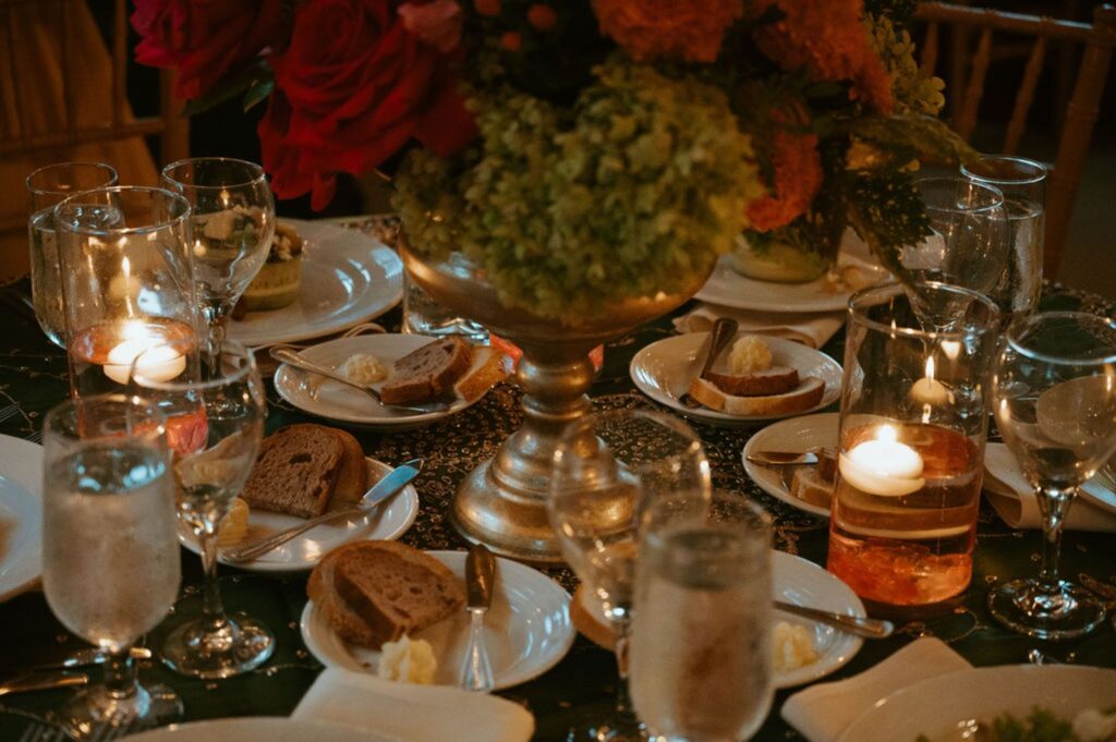 A close-up of a dinner table at a Pace House wedding reception, set with plates of bread and butter, water glasses, wine glasses, and a vibrant flower centerpiece. Lit candles in glass holders add a warm glow to the elegant setting.
