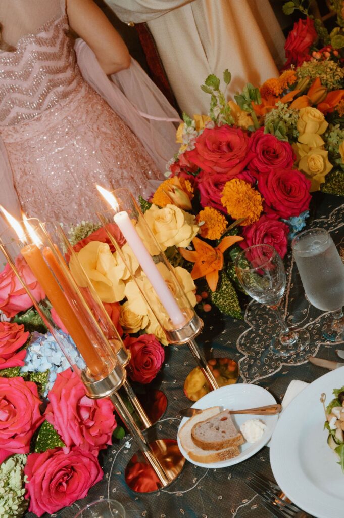 A close-up of a decorated table at a Pace House wedding reception with colorful roses, yellow flowers, lit candles, plates, bread with butter, and a glass of water. In the background, a person in a pale pink sequined dress is partially visible.