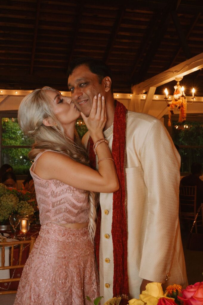 At a Pace House wedding reception, a woman in a pink dress kisses a man wearing a cream-colored sherwani and red scarf on the cheek, surrounded by string lights and flowers in the festive indoor setting.