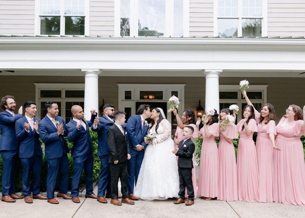 A bride and groom kiss in the center of a wedding party during a progressive cocktail reception. Bridesmaids in pink dresses and groomsmen in blue suits cheer, holding bouquets in front of a white house with columns in Atlanta.