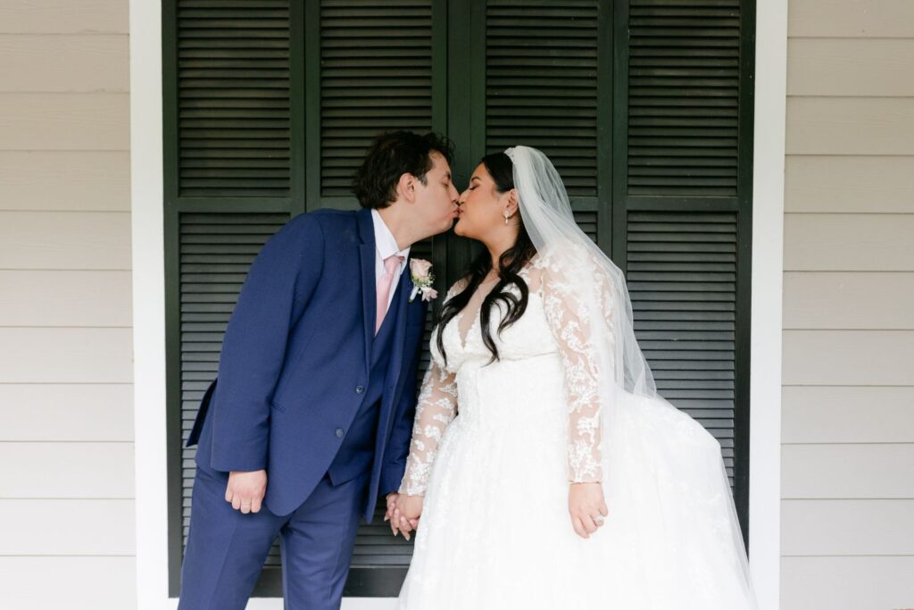A bride and groom holding hands and kissing in front of black window shutters, just after their progressive cocktail reception Atlanta. The groom wears a blue suit, and the bride wears a white lace wedding dress with a veil.