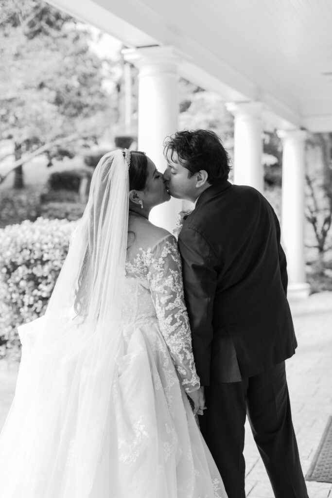 A bride and groom share a kiss outdoors under a covered walkway during their progressive cocktail reception in Atlanta. The bride wears a long-sleeved lace wedding gown with a veil, and the groom is in a dark suit. The scene is captured in black and white.