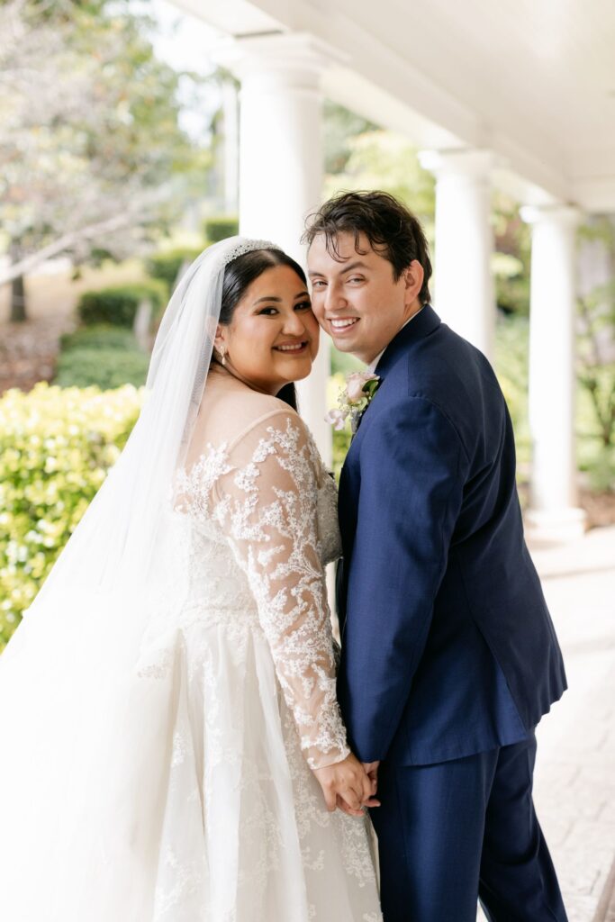 A bride in a white lace gown and veil holds hands with a groom in a blue suit. They smile back at the camera under a covered outdoor walkway, enjoying their progressive cocktail reception in Atlanta amidst white columns and lush greenery.