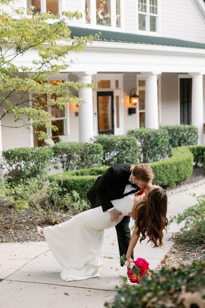 A groom in a black tuxedo dips and kisses a bride in a white gown holding red flowers, in front of a white house with columns and greenery—capturing the romance of a classic Chastain Horse Park wedding reception.