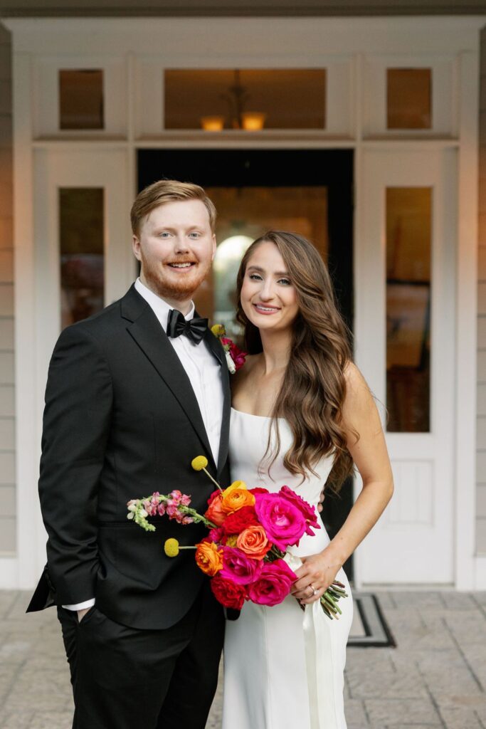 A couple stands smiling in front of a house entrance; the man wears a black suit and bow tie, while the woman in a white dress holds a bouquet of bright pink, orange, and yellow flowers after their Chastain Horse Park wedding reception.