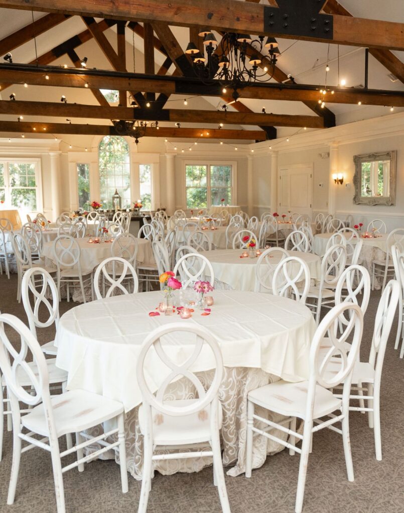 A bright, elegant banquet hall at Chastain Horse Park sets the scene for a wedding reception, with round tables draped in white cloths, floral centerpieces, and string lights. Wooden ceiling beams and large windows highlight the inviting space.