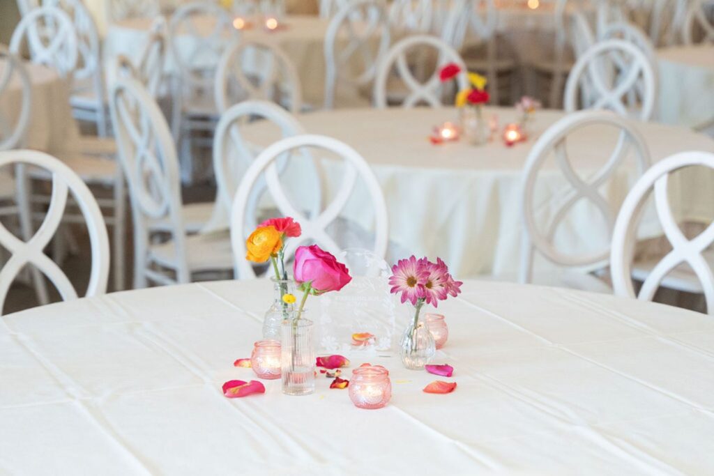 A round table with a white tablecloth is decorated with small vases of colorful flowers, rose petals, and votive candles. Similar tables fill the background, creating an elegant Chastain Horse Park wedding reception setting.