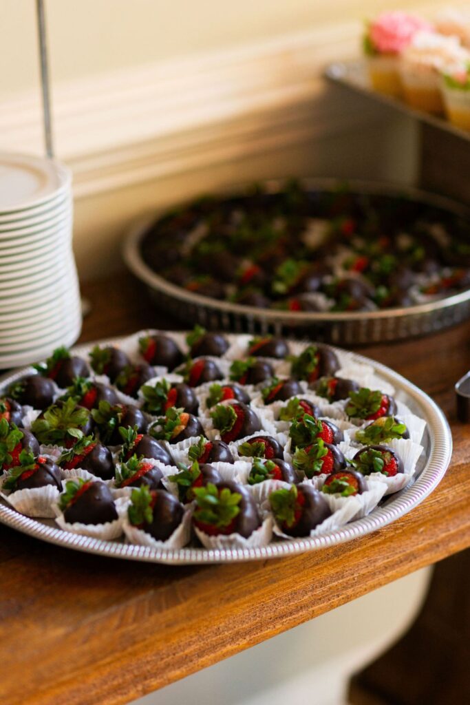 At a Chastain Horse Park wedding reception, a silver tray filled with chocolate-covered strawberries in white paper cups sits on a wooden table, with another tray of similar treats and stacked plates in the background.
