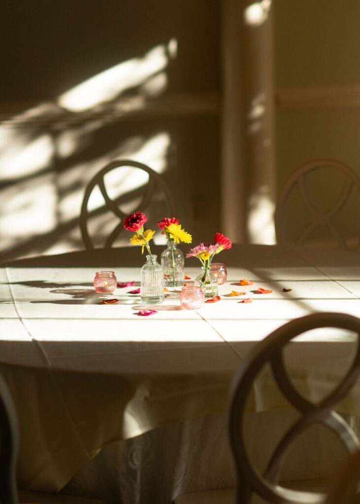 At a Chastain Horse Park wedding reception, a round table is decorated with small vases of red and yellow flowers, scattered petals, and pink glass candle holders. Sunlight casts shadows and light across the table and nearby chairs.