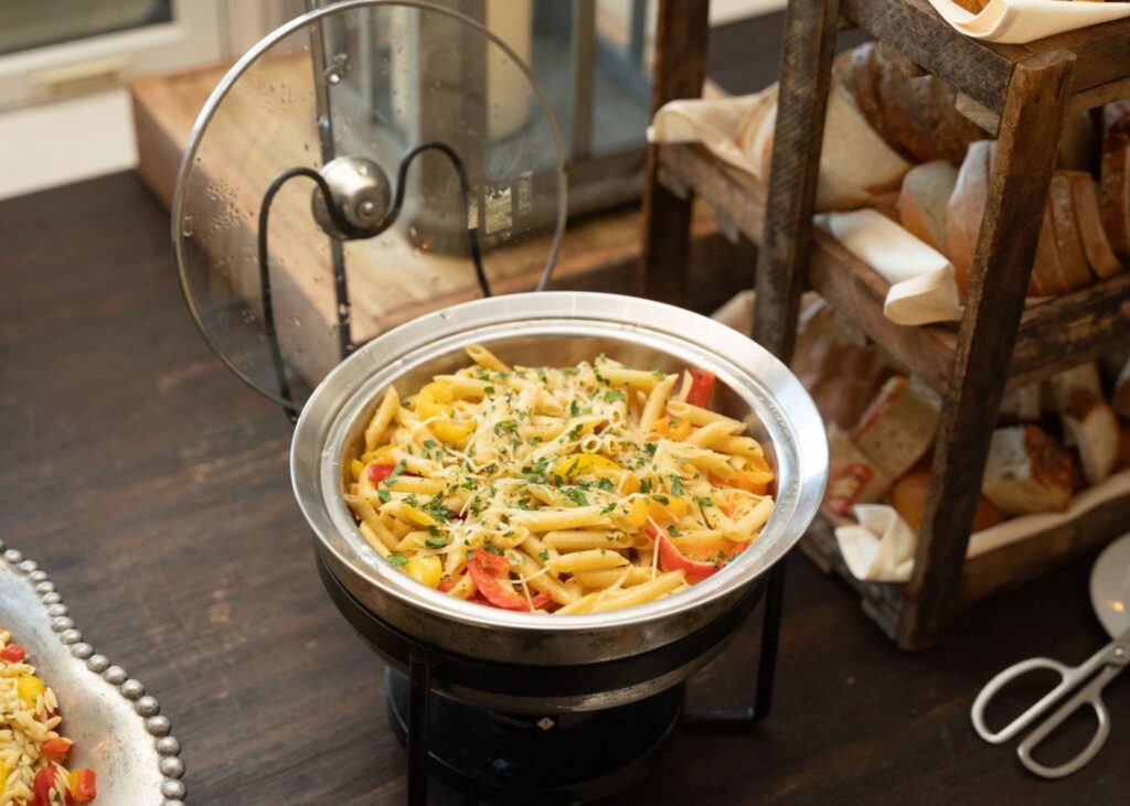 A buffet setup at a Chastain Horse Park wedding reception features a metal chafing dish filled with penne pasta mixed with bell peppers and herbs, set beside a wooden rack stocked with assorted bread.
