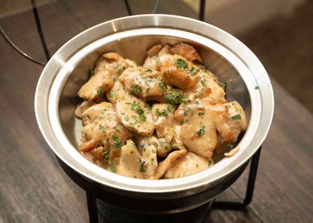 A stainless steel bowl filled with creamy, sauced chicken pieces, topped with chopped herbs, rests on a black metal stand on a wooden surface—perfect for serving at a Chastain Horse Park wedding reception.
