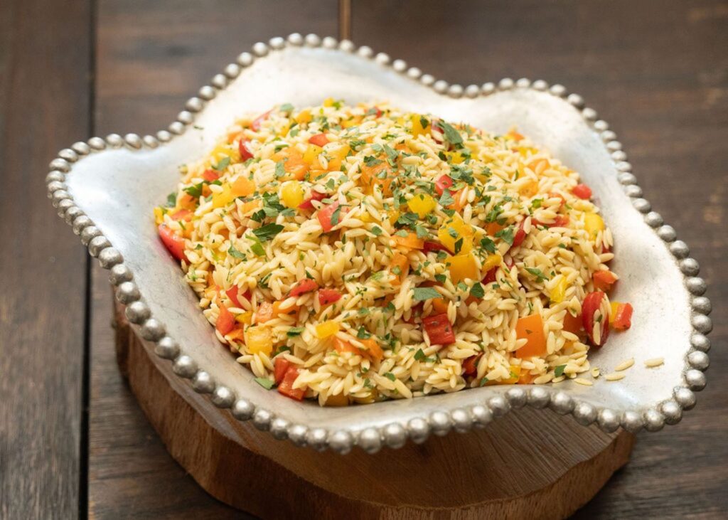 A decorative silver bowl holds orzo pasta mixed with diced red and yellow bell peppers and chopped parsley, perfect for serving at a Chastain Horse Park wedding reception, placed elegantly on a wooden surface.