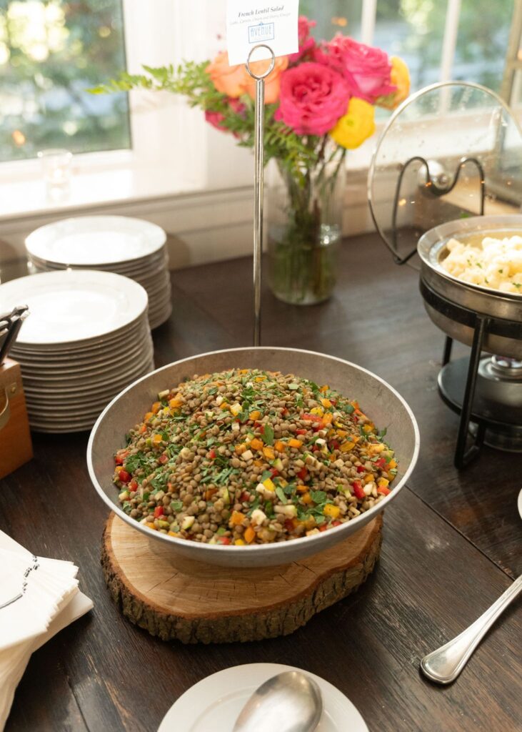 A large bowl of fresh lentil salad with chopped vegetables is displayed on a wooden slab at a Chastain Horse Park wedding reception. Plates are stacked nearby, with flowers and other dishes in the background on a wooden table.