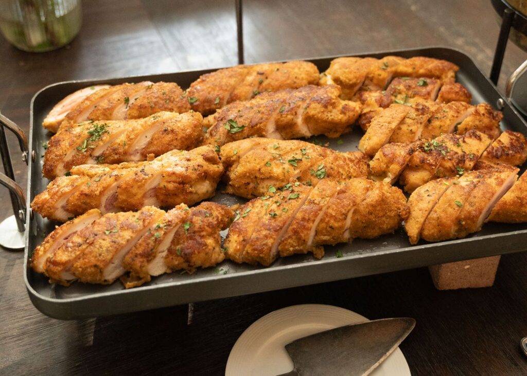 At a Chastain Horse Park wedding reception, a serving tray filled with golden-brown breaded chicken, garnished with fresh herbs, sits on a dark wooden table. A plate and serving utensil are placed nearby for guests to enjoy.