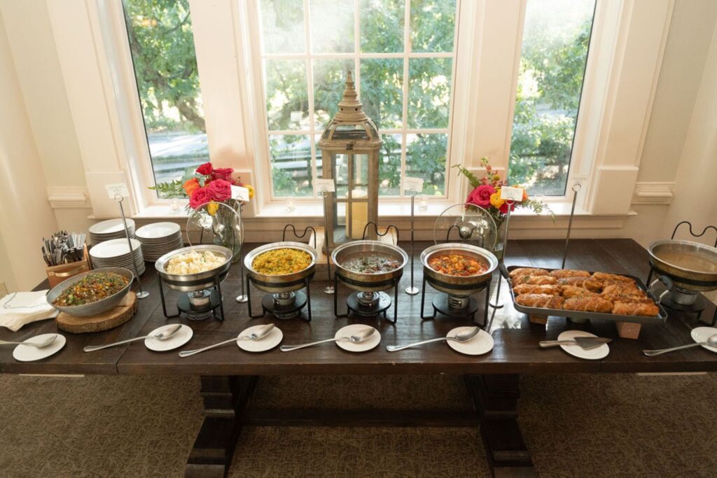 At a Chastain Horse Park wedding reception, a buffet table is set with chafing dishes filled with hot foods, plates, and utensils. Red flowers and lanterns decorate the space, while large windows fill the room with natural light.