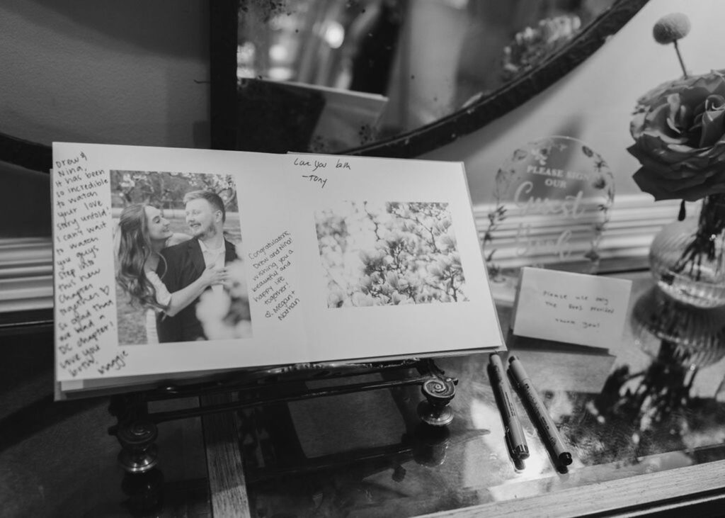 A black-and-white photo of an open guest book at a Chastain Horse Park wedding reception, featuring handwritten notes, a couple's photo, pens, a bouquet of flowers, and a sign inviting guests to sign, all reflected in a mirror.