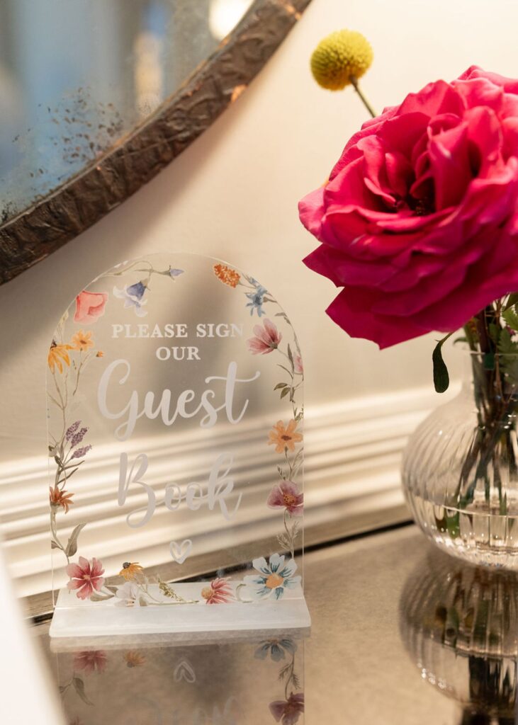 A clear acrylic sign with floral decorations reads Please sign our Guest Book beside a bright pink rose in a ribbed glass vase, placed on a reflective surface at a Chastain Horse Park wedding reception.