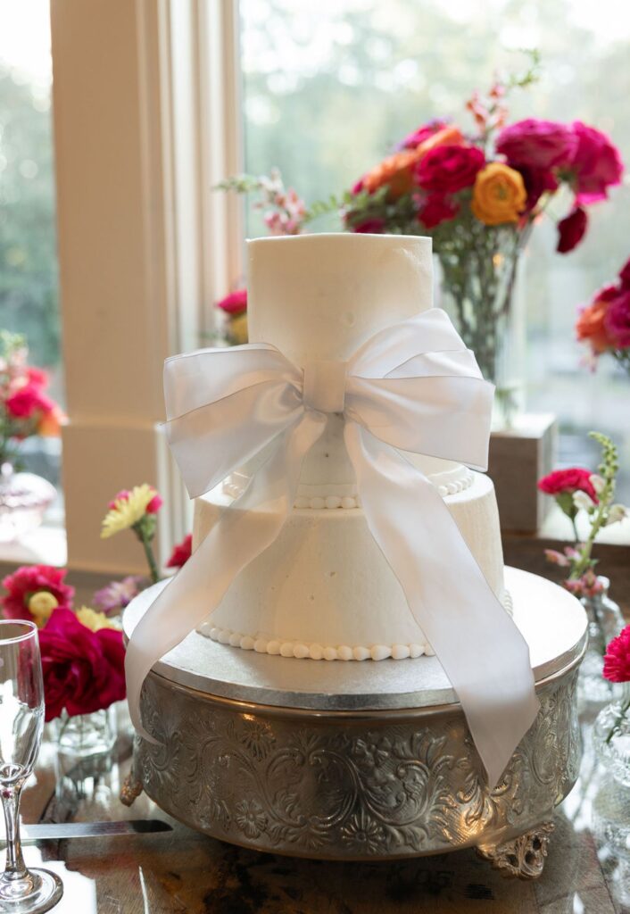 At a Chastain Horse Park wedding reception, a three-tier white wedding cake adorned with a large satin bow sits on an ornate silver stand, surrounded by red and pink flowers near a sunlit window.