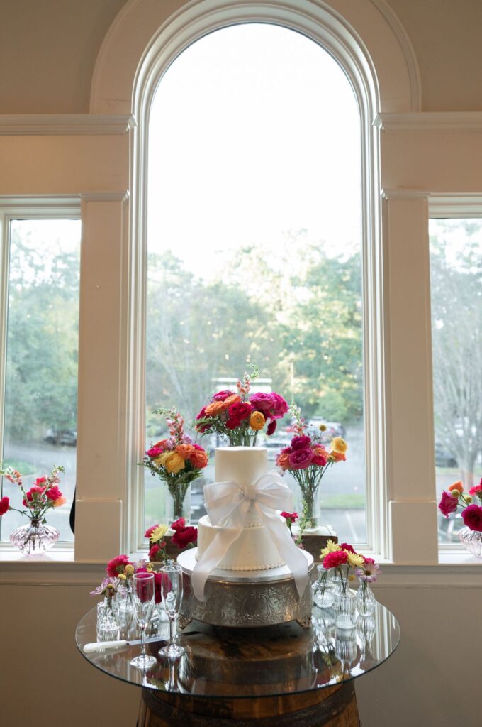 A white wedding cake with a large bow and bright flower arrangement sits on a glass table at a Chastain Horse Park wedding reception, surrounded by colorful floral bouquets and glasses, in front of tall arched windows overlooking greenery.